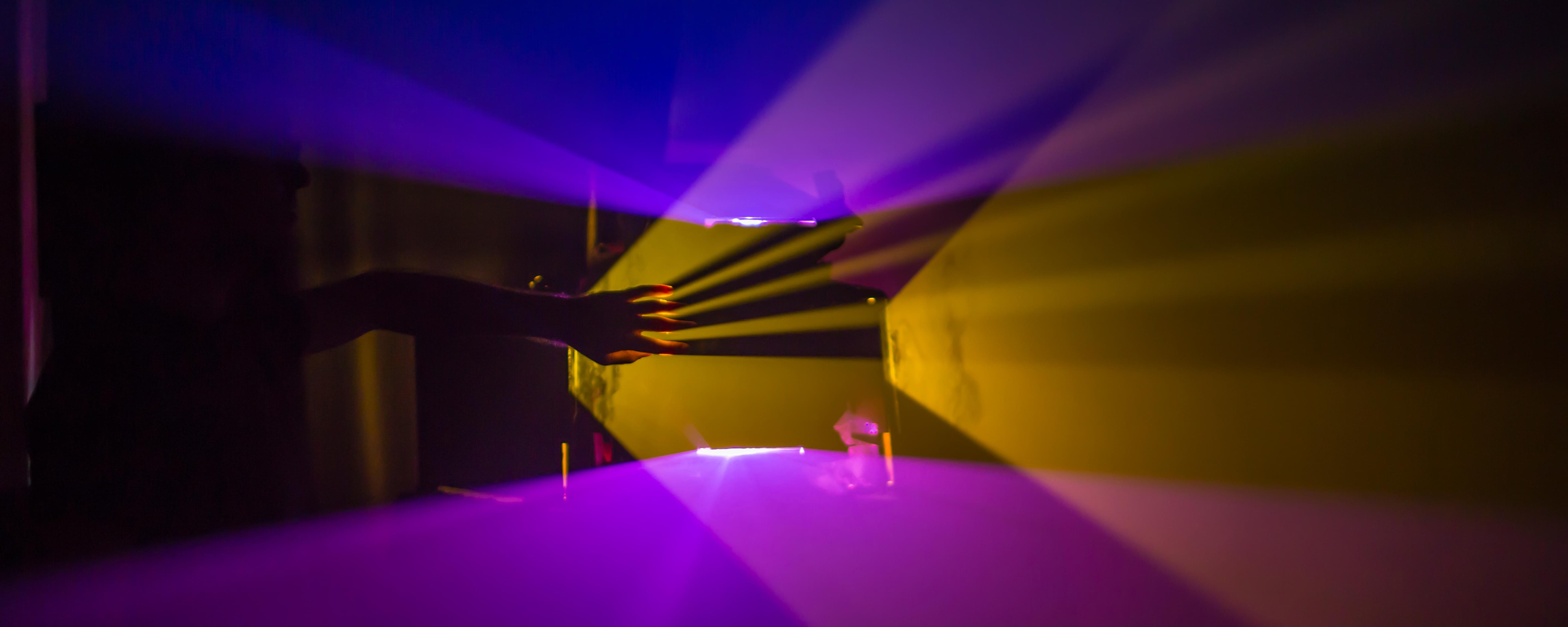 Photograph of dark room with planes of yellow and purple light emminating from projectors. A hand is in one of the planes creating a shadow lake of fingers.