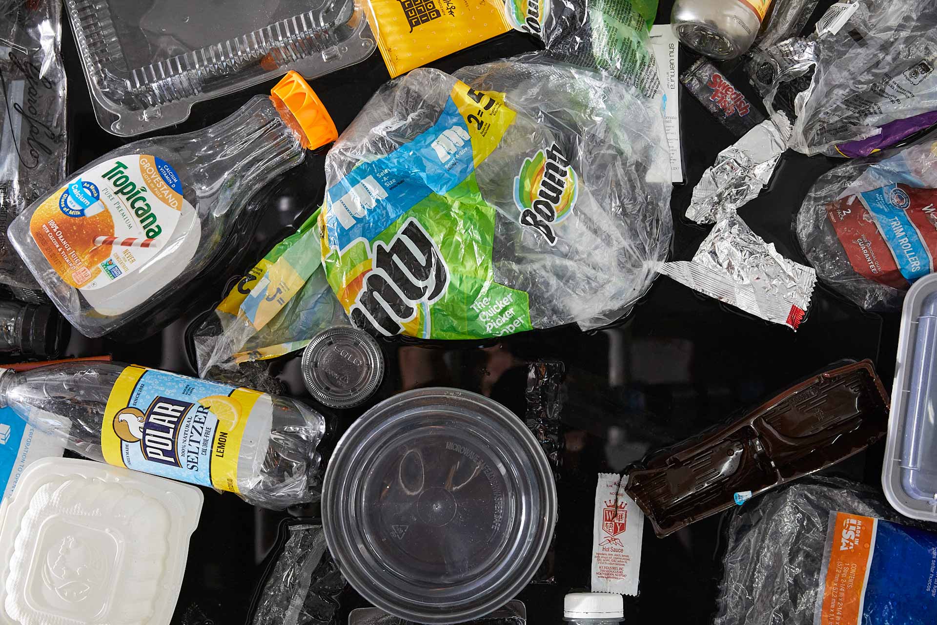 Tight close-up photograph of about 20 single-use plastic items jumbled together. Most prominently featured is a Bounty plastic package wrapping, a Tropican organge juice bottle, and a circular take out container.