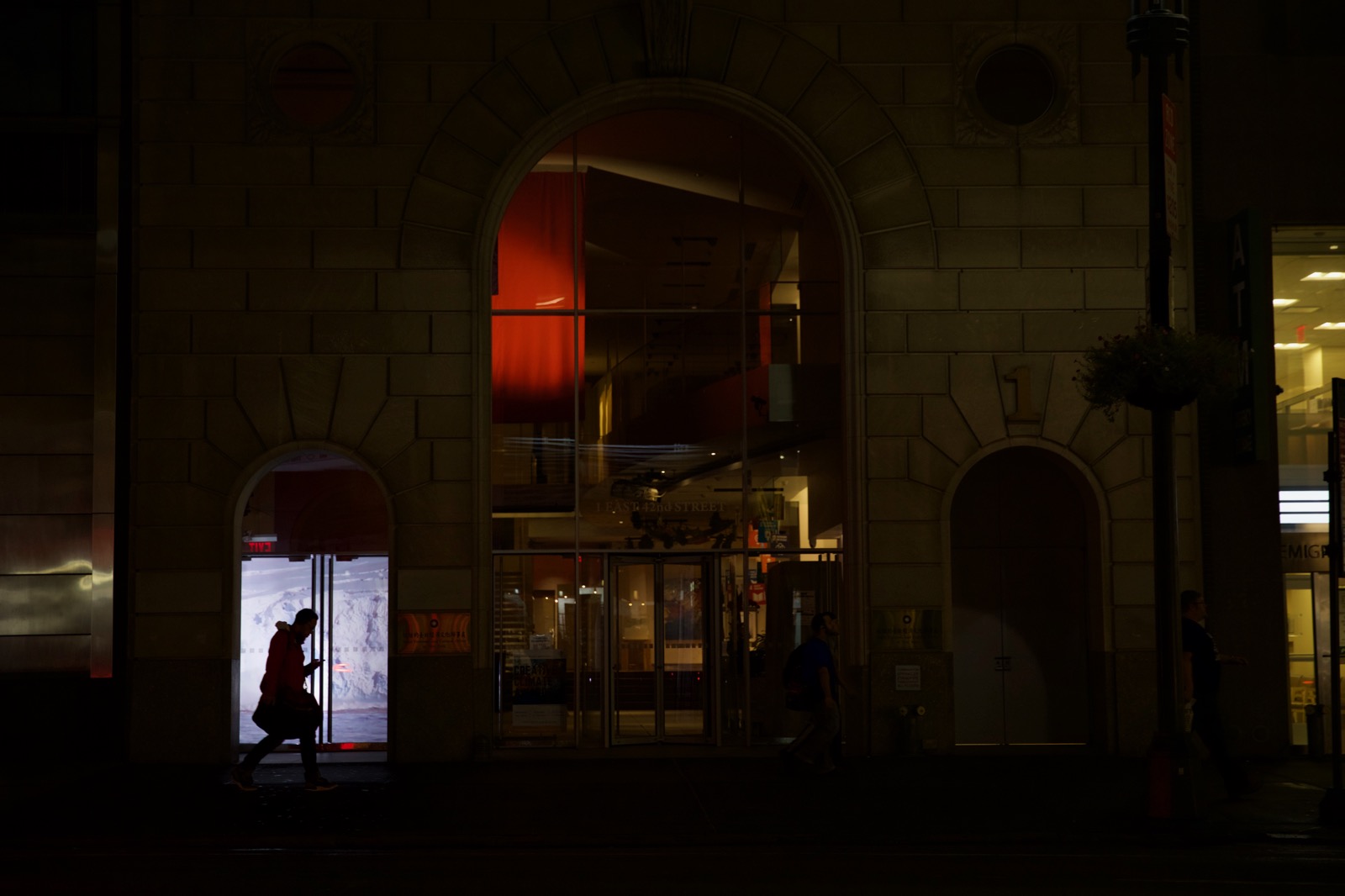 Very dark nighttime photograph of a silhouetted figure walking down the street looking at their phone. Behind him is a projected image of glaciers in a small storefront window.