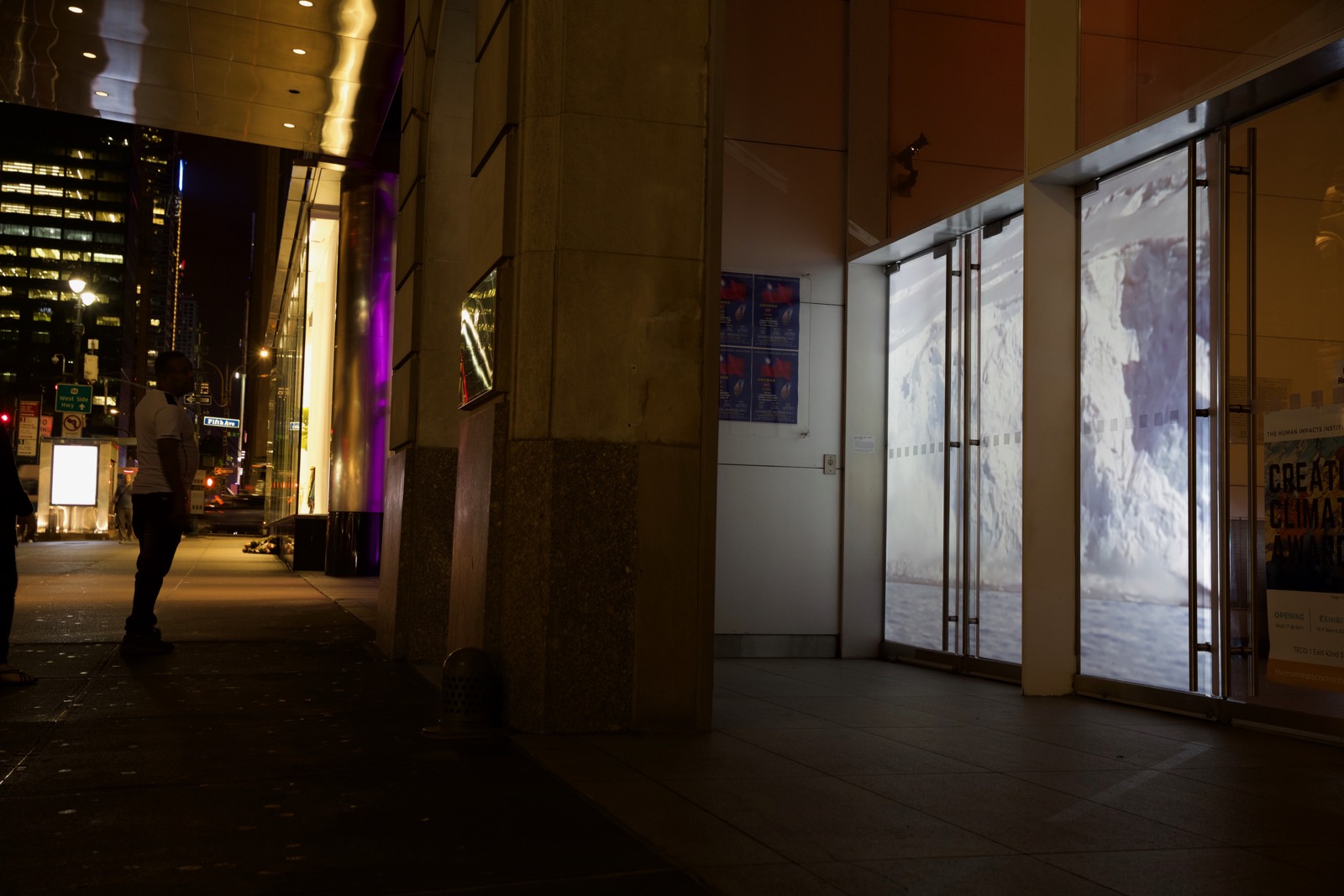 Nighttime photograph of a city street—a street sign in the far background says 'Fifth Avenue.' On the left side is a passerby who has stopped and is looking at the projection of an arctic scene (glaciers) in the windows of the building on the right.