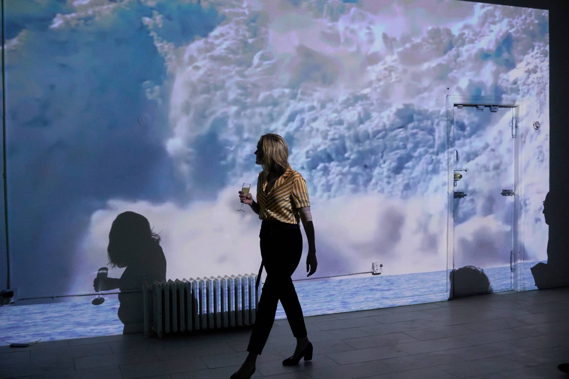Slighly askew photograph of a women in cocktail attire, holding a cocktail, walking in front of a projection of a glacier collapsing