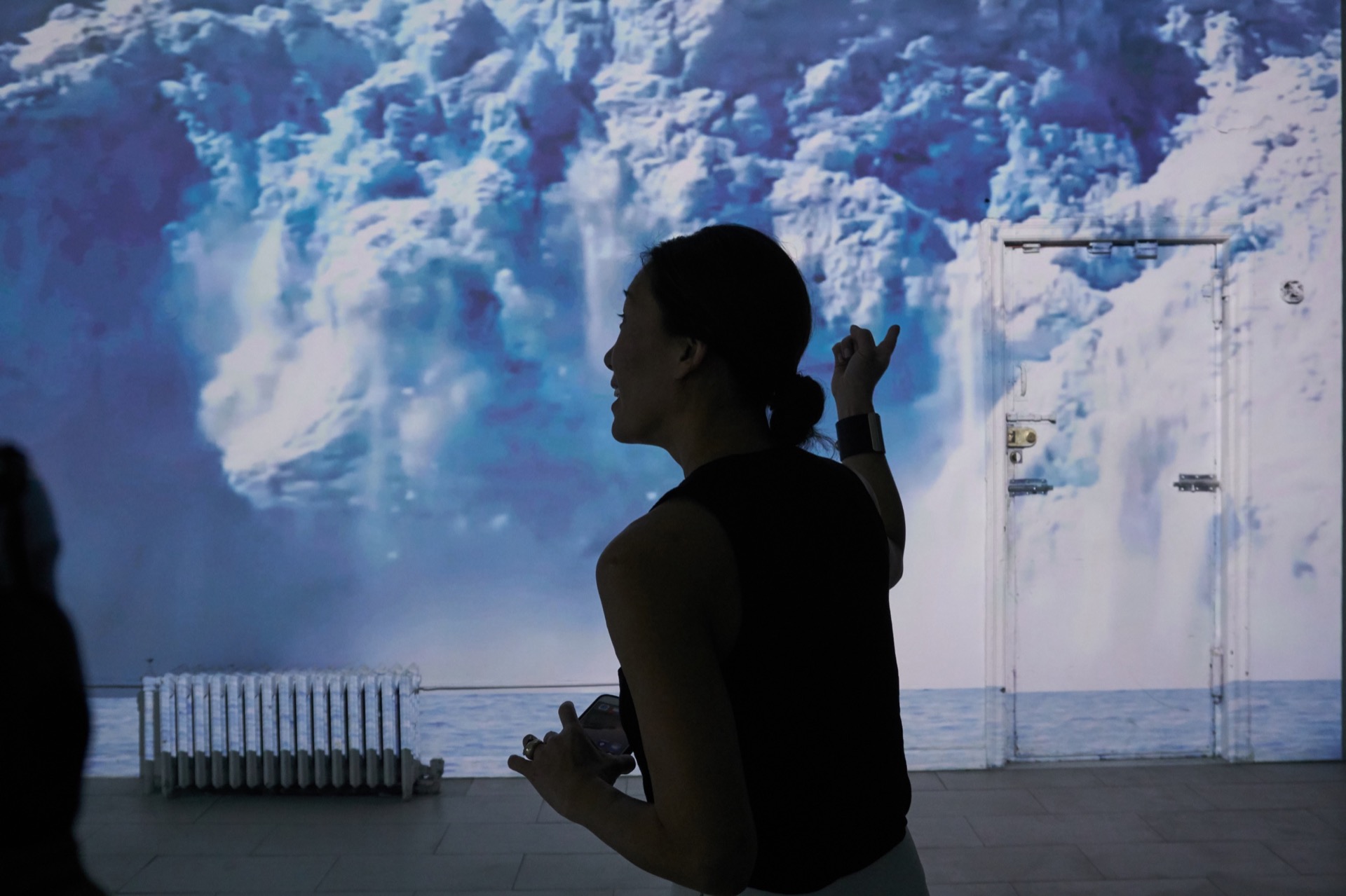 Close-up photograph of a person's head, in 3/4 profile in the middle of the frame. She is pointing to the wall behind her, onto which is projected an image of a glacier collapsing. She appears to be saying something.