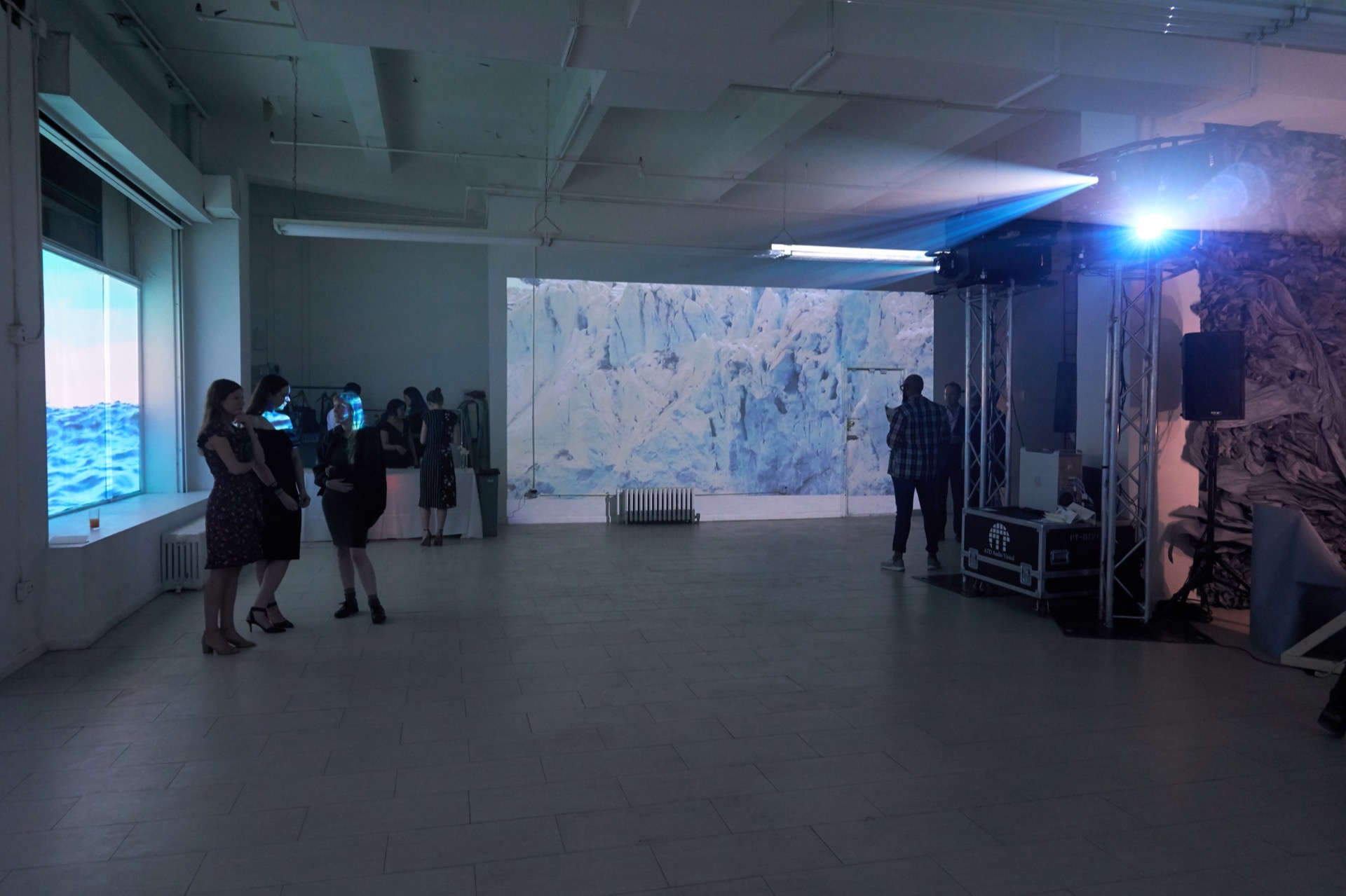 Wide shot of the inside of a gallery showing two different projected works. On the left, an image of a body of water is projected into the gallery windows. Straight back, an image of an Arctic scene—close up of a glacier—is projected. There are about a dozen people in cocktail attire milling about. On the right, a two sharply bright lights are emanating from the lens of large projectors.